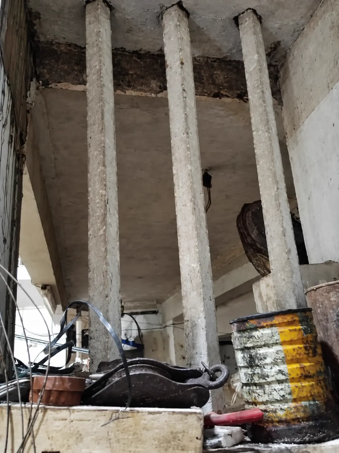 三根混凝土方柱撑着一层加建的楼板，地面堆着杂物与油漆桶