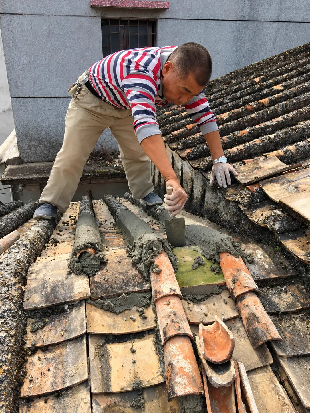 一位师傅蹲在旧瓦屋顶上，用灰匙把灰浆抹进瓦垄之间，屋顶瓦片已经斑驳